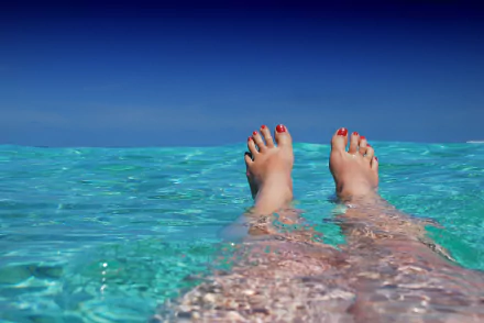 A serene image of a woman's feet submerged in clear, turquoise pool water, with blue skies above, creating a tranquil and refreshing atmosphere.