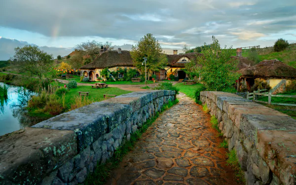 A vibrant HDR image of Hobbiton village featuring a stone bridge over a calm stream and a cobblestone path leading through the man-made scenic landscape.
