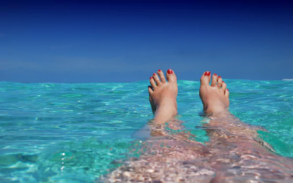 A serene image of a woman's feet submerged in clear, turquoise pool water, with blue skies above, creating a tranquil and refreshing atmosphere.