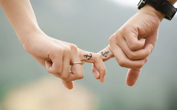A close-up of a couple's hands linking their fingers, showcasing a tattoo and a ring, symbolizing love. The soft background enhances this intimate moment, creating a striking HD wallpaper.