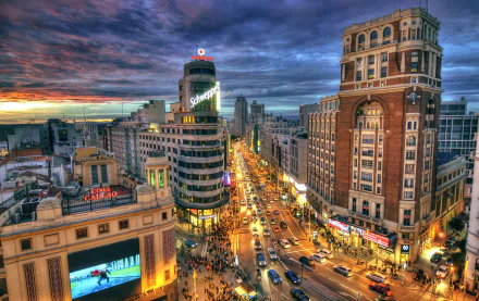 Evening cityscape of Madrid, Spain, showcasing busy roads, illuminated buildings, cars, and people against a vibrant, colorful sky in this HD desktop wallpaper.