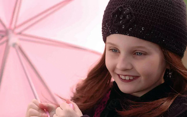 A young girl smiles while holding a pink umbrella, wearing a stylish black hat. This image features Abigail Breslin from the movie The Ultimate Gift, serving as a vibrant HD wallpaper.