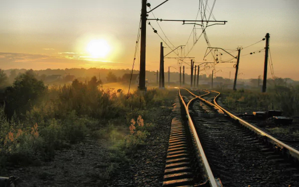 A serene HD wallpaper featuring a winding railroad track under a warm sunrise, framed by tall grasses and electric poles, capturing the essence of man-made beauty in nature.