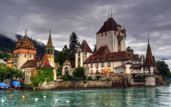 Oberhofen Castle in Switzerland stands majestically by the water under a dramatic cloudy sky, showcasing its historic man-made architecture in vivid detail.
