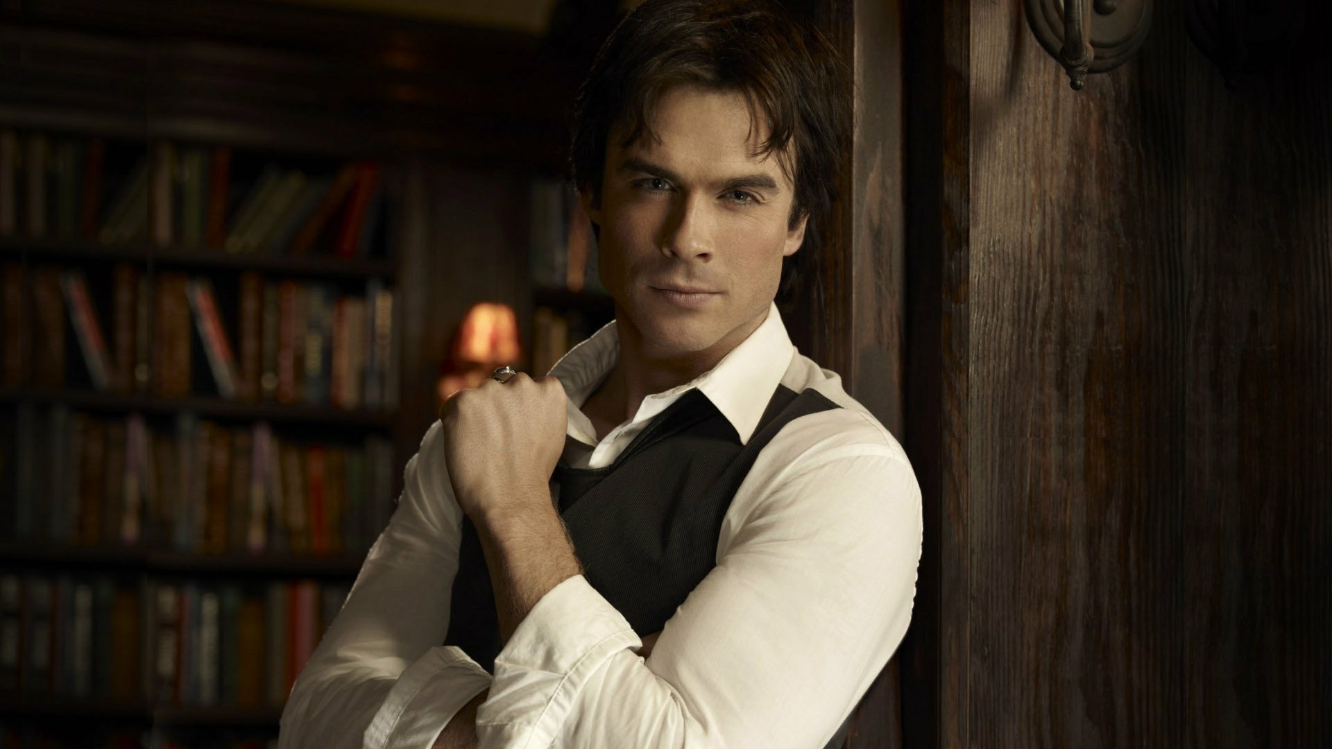 HD desktop wallpaper featuring a character from the TV show The Vampire Diaries, standing confidently in a cozy library setting.