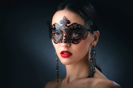 A brunette woman with striking green eyes and bold red lipstick gazes intently, wearing a decorative black mask and elegant earrings, creating an intriguing photographic portrait.