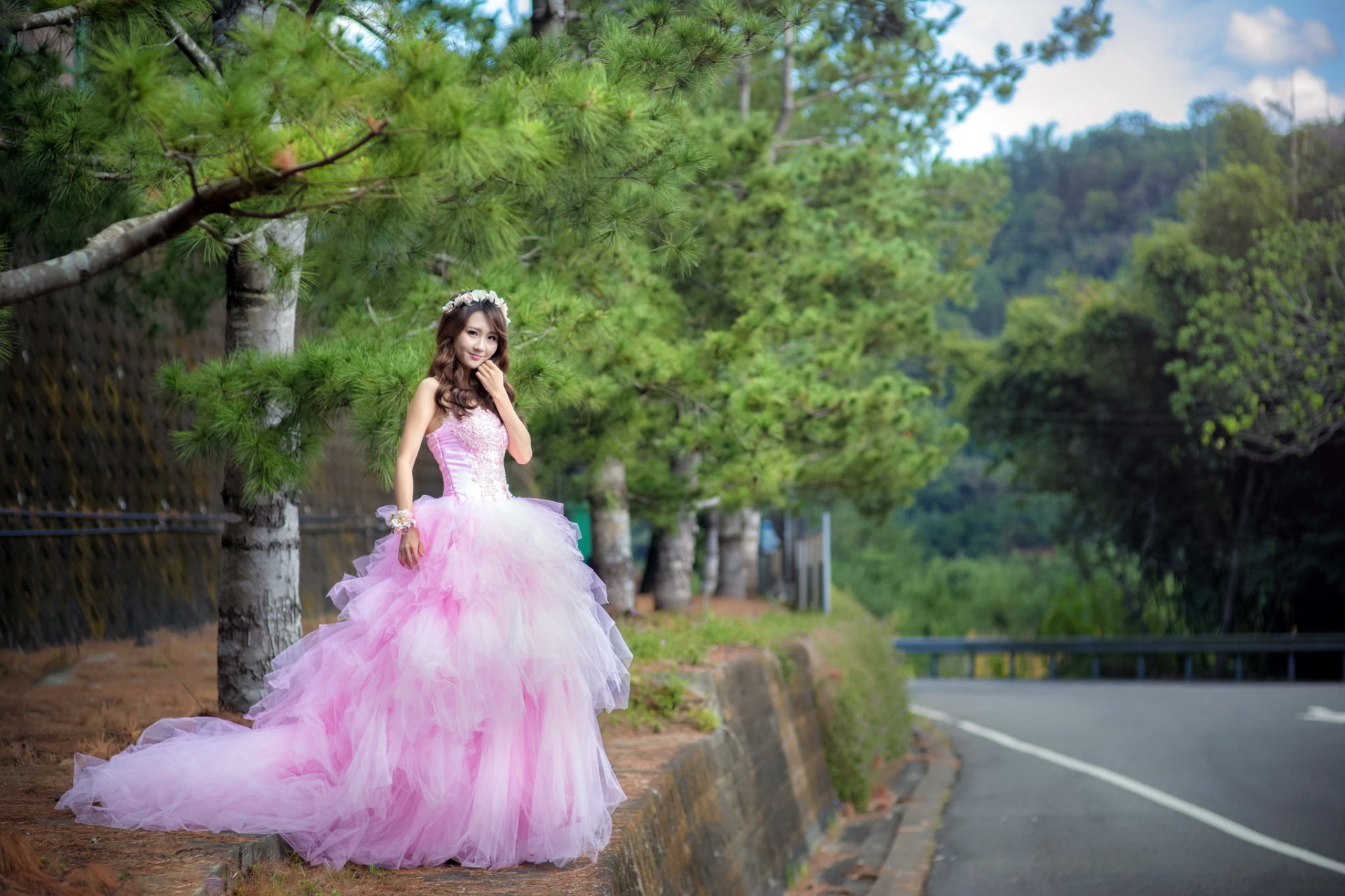 A bride stands outdoors in a lush, green setting, wearing a pink, voluminous wedding dress. The background shows a winding road and dense trees. This HD image is a desktop wallpaper.