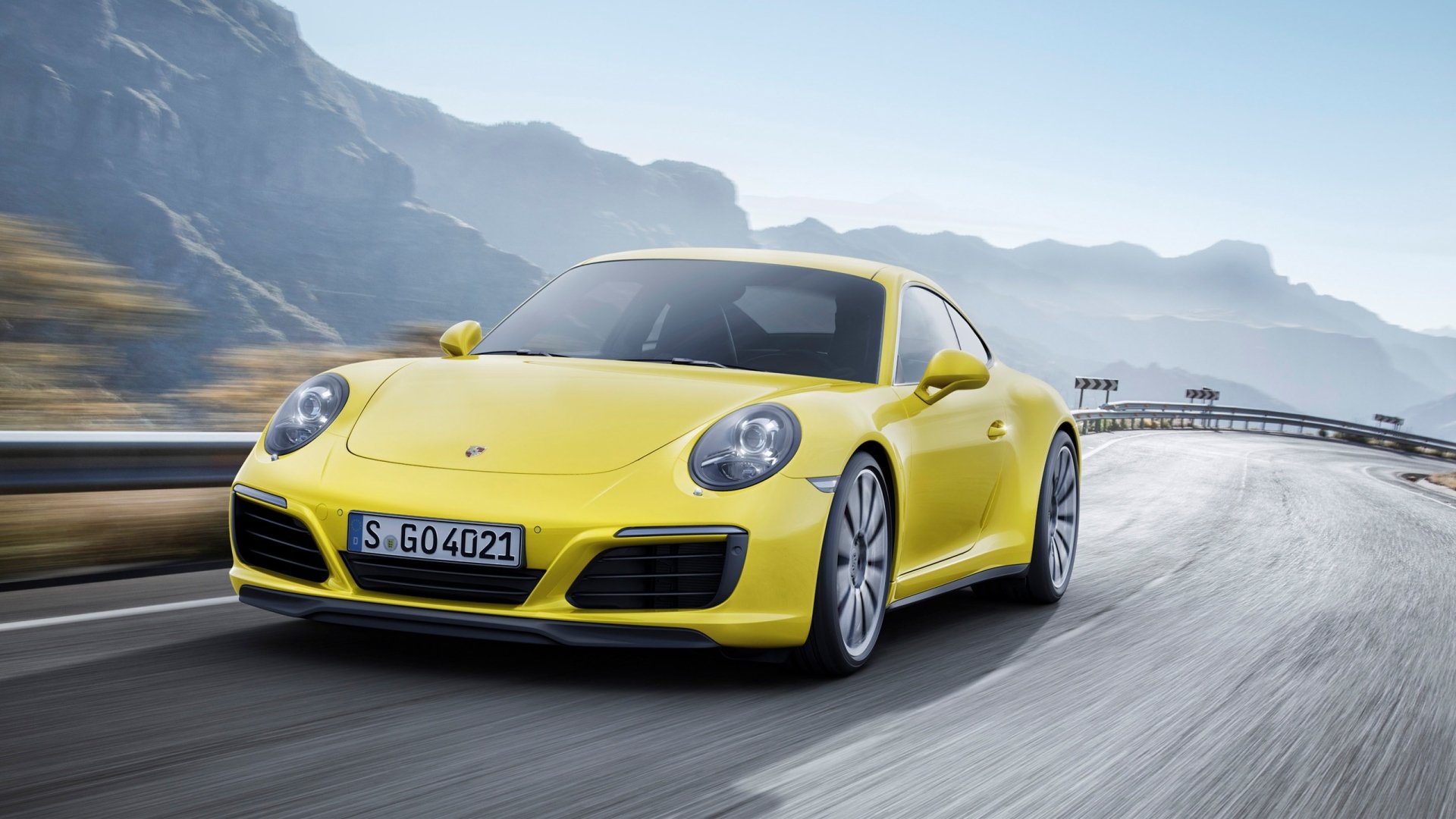 A vibrant yellow Porsche 911 Carrera accelerates along a winding road, surrounded by mountainous scenery, showcasing the vehicle's sleek design and dynamic presence.