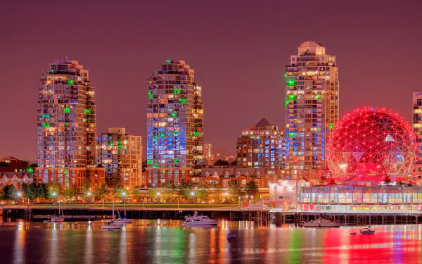 A vibrant night cityscape of Vancouver, showcasing illuminated high-rise buildings and the iconic red dome, reflecting beautifully on the water.