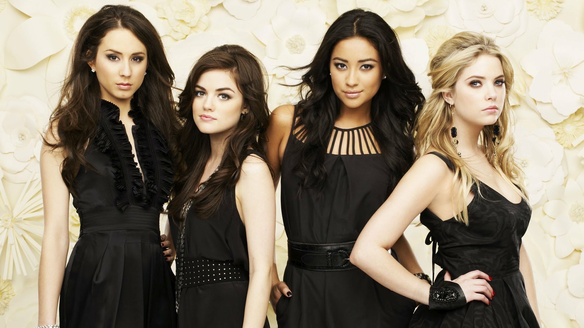 Four main characters from the TV show Pretty Little Liars pose together in stylish black outfits against a textured background, showcasing their signature friendship and intrigue.
