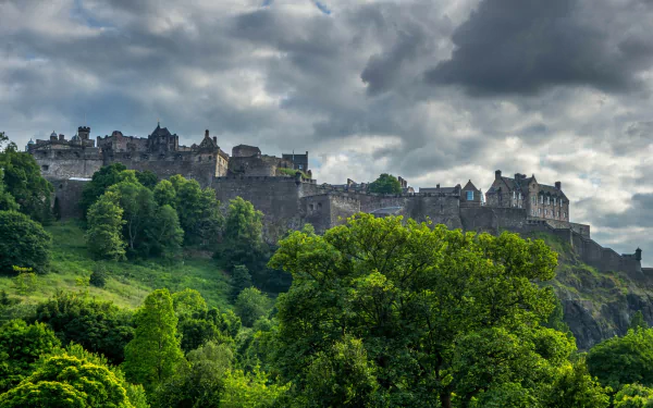 2K Quad HD PC desktop wallpaper: Edinburgh Castle perched on a rocky crag under brooding clouds, verdant trees framing the man-made fortress.