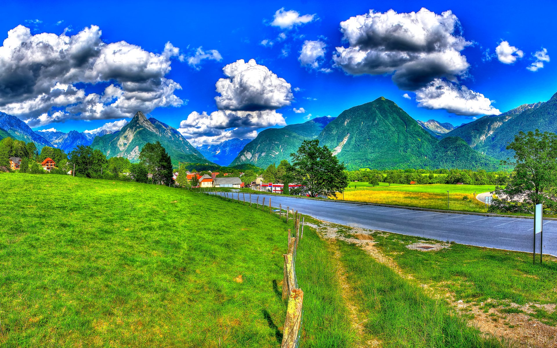 HD landscape photography of a vibrant Slovenian village with green grass, winding road, dramatic clouds, and towering mountains under a bright blue sky.
