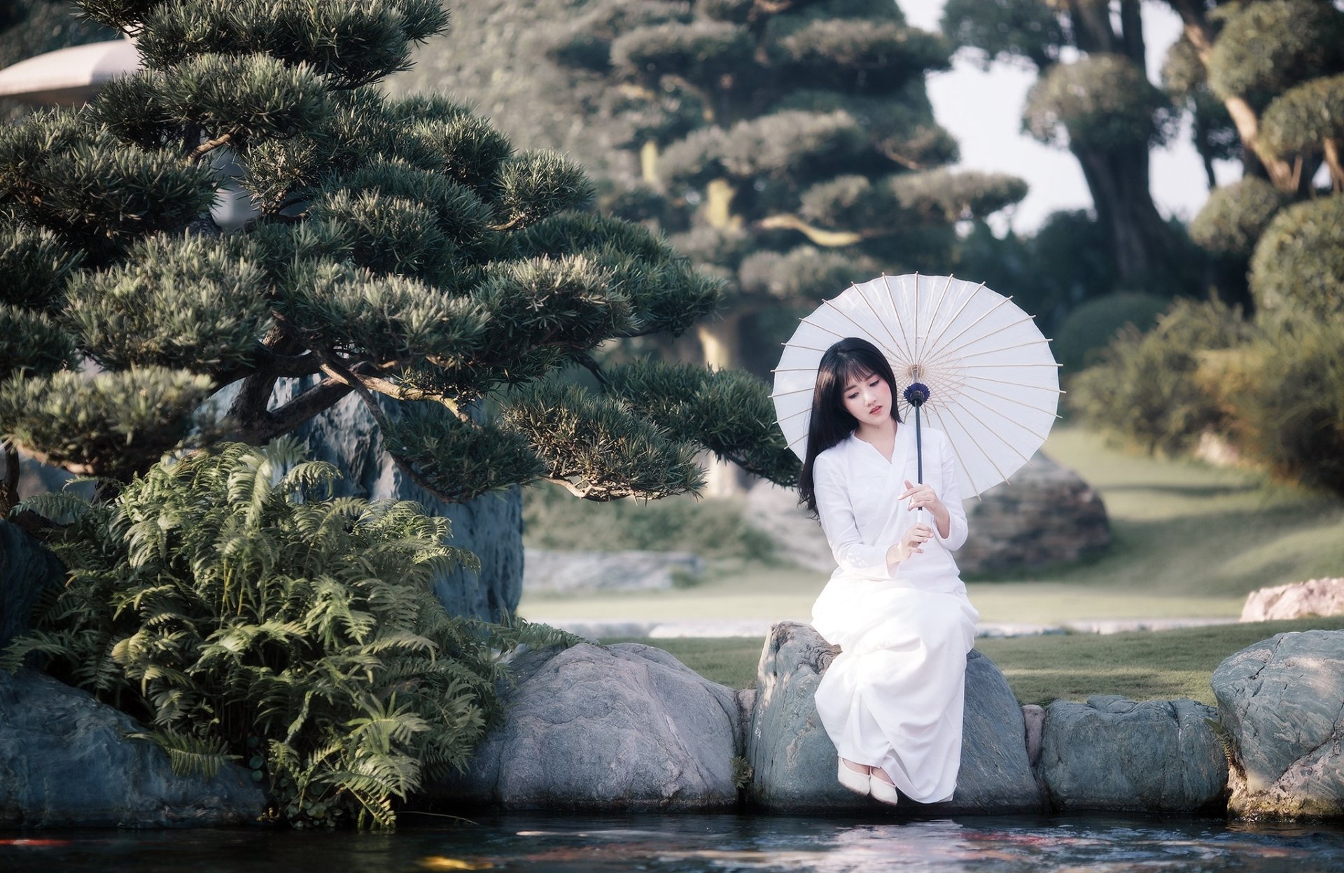 A brunette Asian woman sits outdoors by a tranquil garden pond, holding a white umbrella, surrounded by lush greenery and elegant trees, creating a serene and picturesque scene.