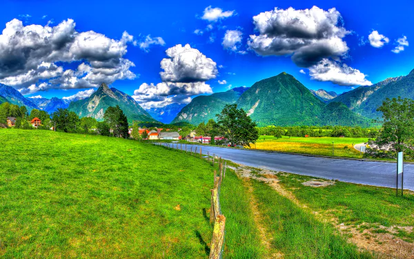 HD landscape photography of a vibrant Slovenian village with green grass, winding road, dramatic clouds, and towering mountains under a bright blue sky.