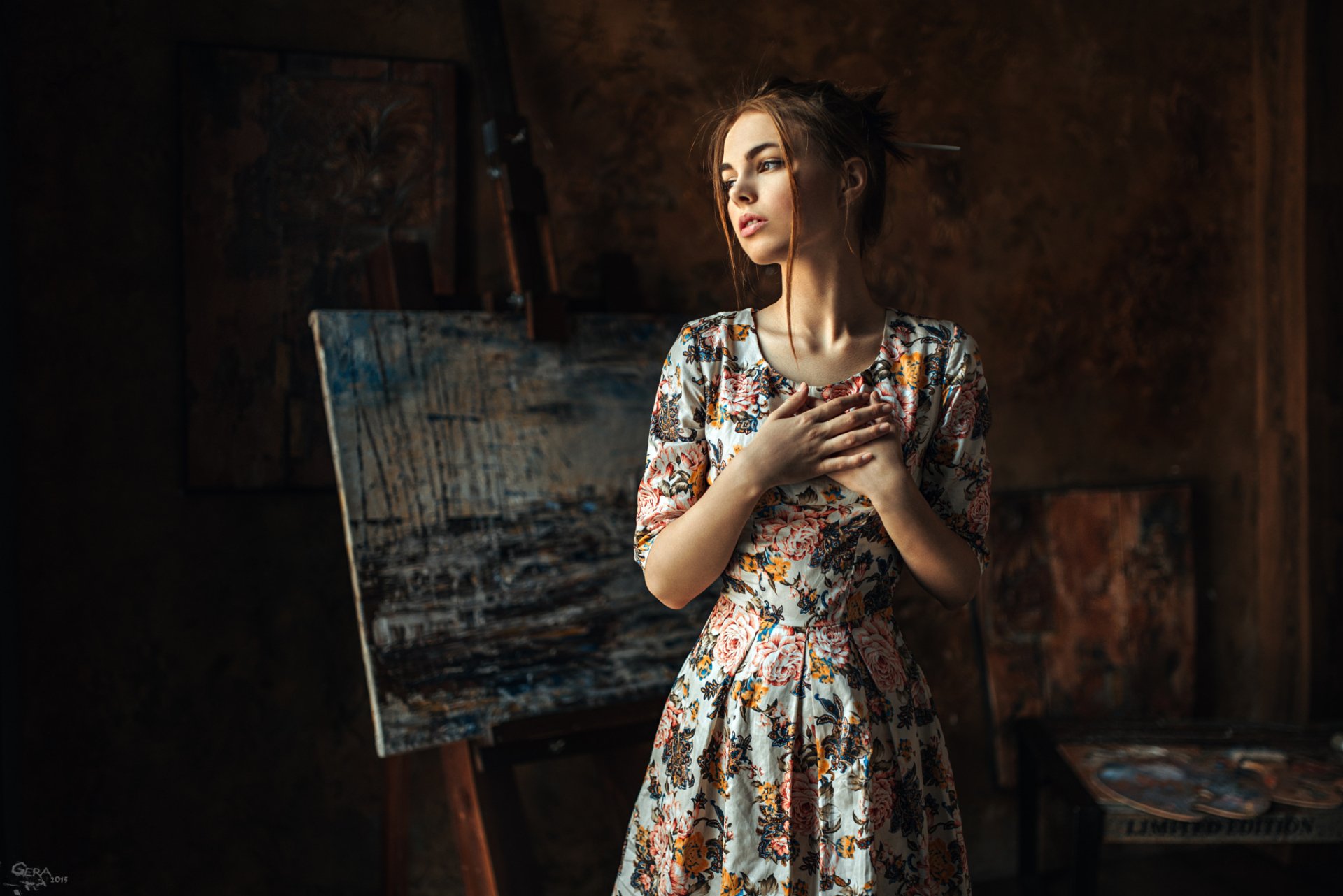 HD desktop wallpaper featuring a brunette woman with brown eyes in a floral dress, standing in front of paintings with a thoughtful expression, creating a serene, artistic mood.