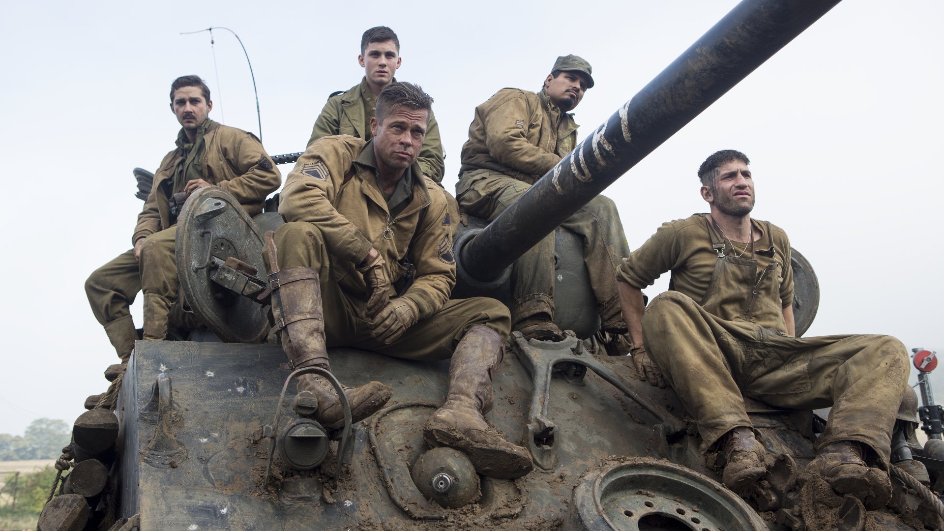 A gripping scene from the movie Fury, featuring Shia LaBeouf, Brad Pitt, Logan Lerman, Jon Bernthal, and Michael Peña, captured atop a tank in muddy military attire.
