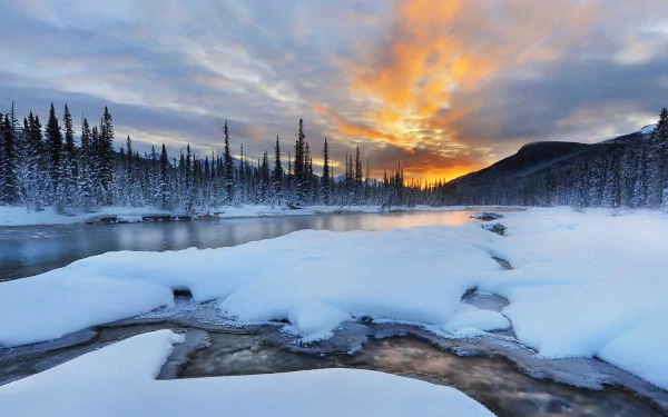 Snow-covered river flows through a forested landscape with a vibrant sunset sky and mountain backdrop in Banff National Park, Alberta, Canada during winter.