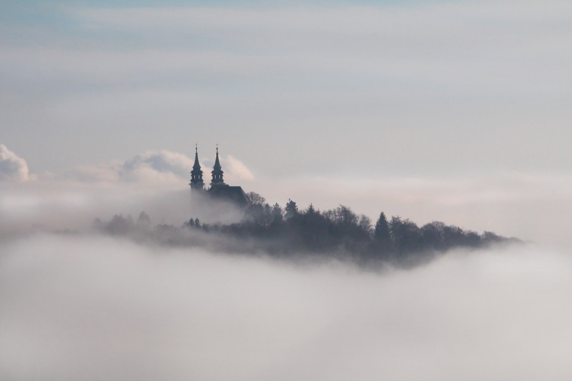 HD PC desktop wallpaper of a white fog enveloping a religious church with twin spires, surrounded by trees, creating a serene and mystical atmosphere.
