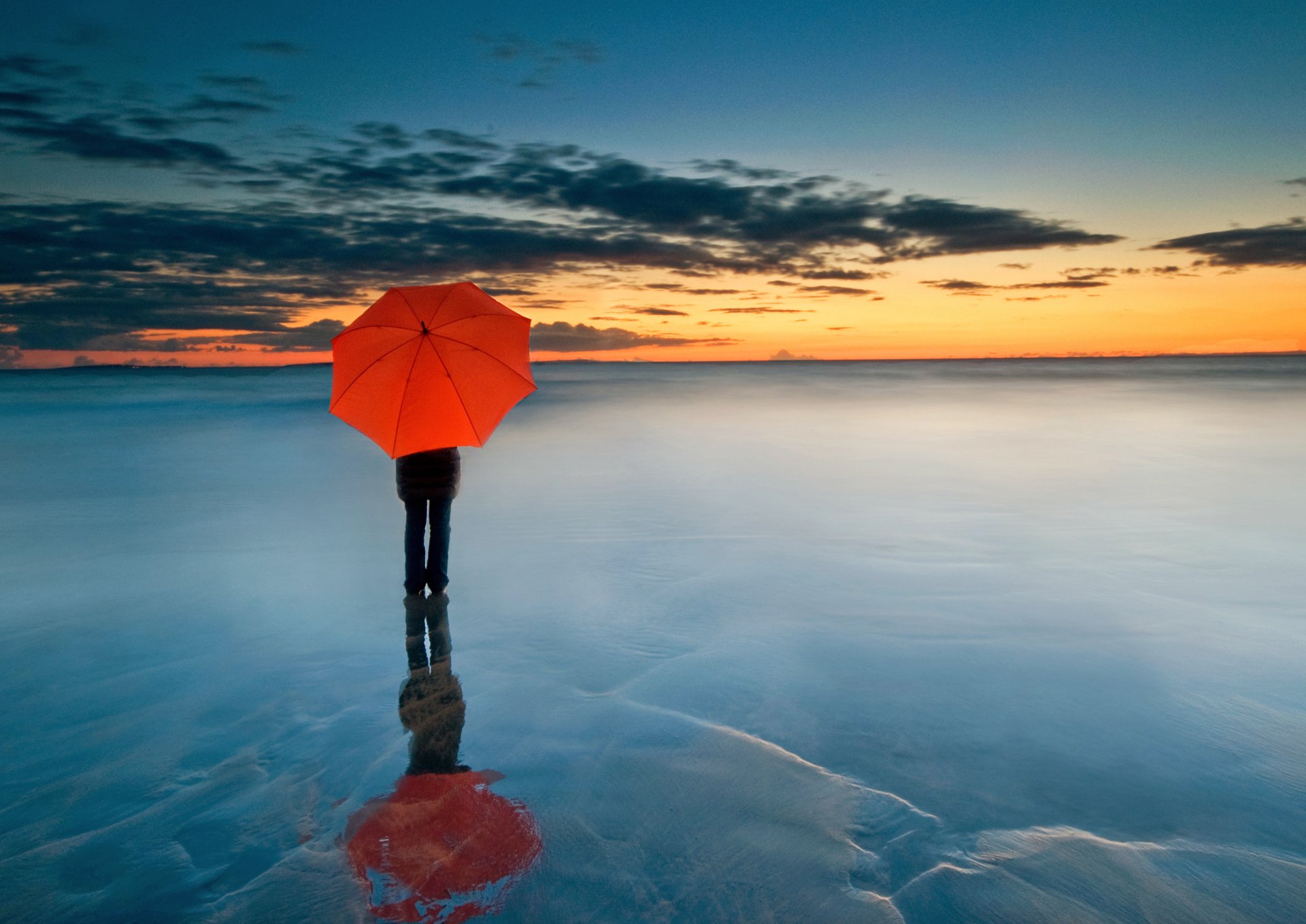 A lone figure with a red umbrella stands on a frozen sea at sunset, reflecting on the icy surface beneath a dramatic cloud-filled horizon in this 4K Ultra HD scenic wallpaper.