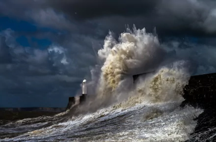 A dramatic ocean storm with towering waves crashing against a man-made structure and a lighthouse in the background, set against a gray, cloud-filled sky. HD desktop wallpaper.