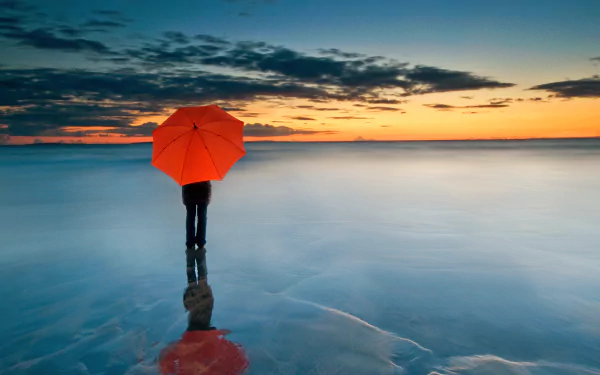 A lone figure with a red umbrella stands on a frozen sea at sunset, reflecting on the icy surface beneath a dramatic cloud-filled horizon in this 4K Ultra HD scenic wallpaper.