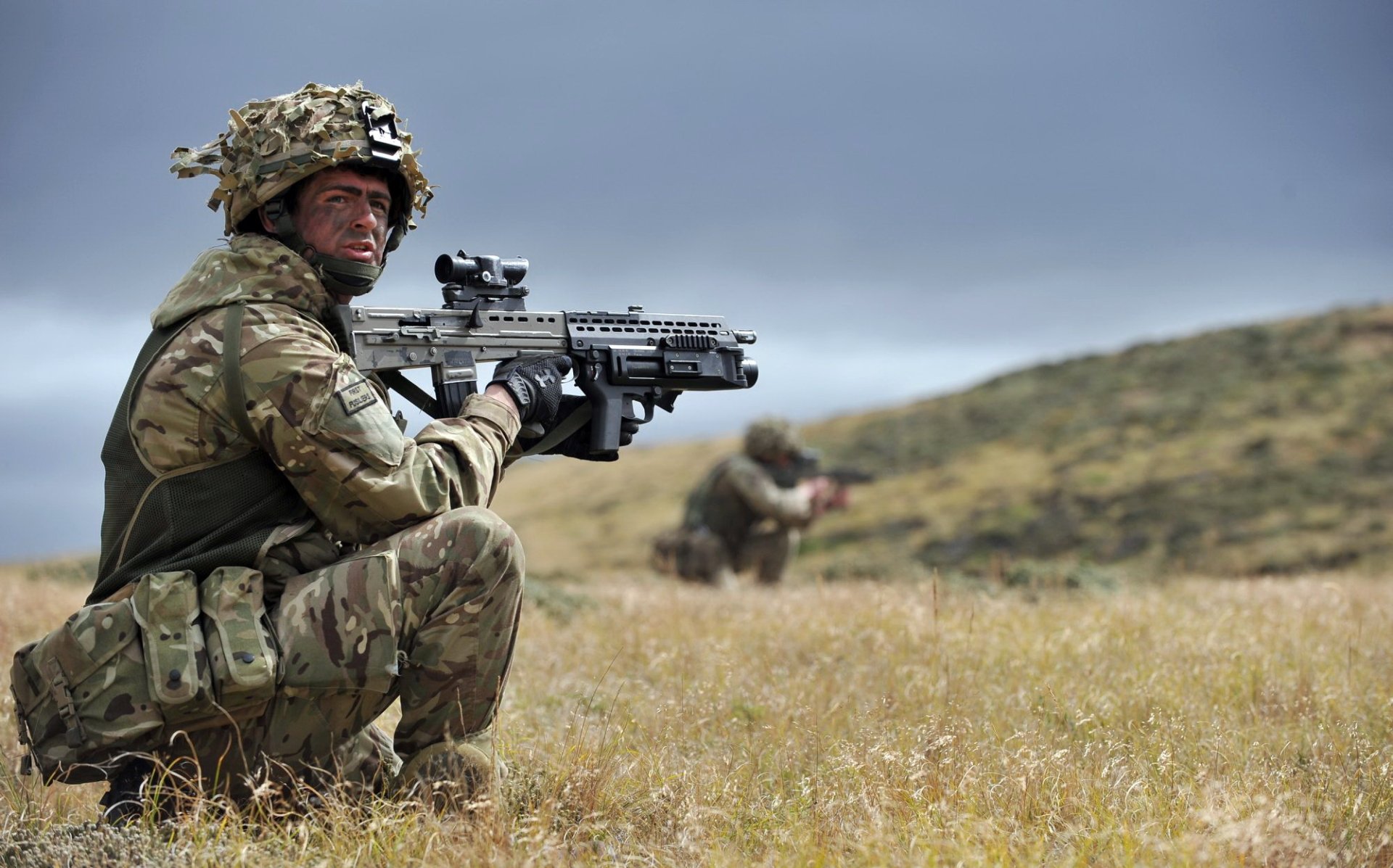 HD PC desktop wallpaper featuring a soldier in military gear aiming an assault rifle, with another soldier blurred in the bokeh background in a grassy field.