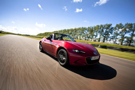 HD desktop wallpaper featuring a red Mazda MX-5 driving on a sunny road with trees and blue sky in the background.