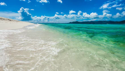 A serene tropical beach scene featuring turquoise waters gently lapping against soft sand, with clouds scattered across a vibrant blue sky and mountains visible on the horizon.