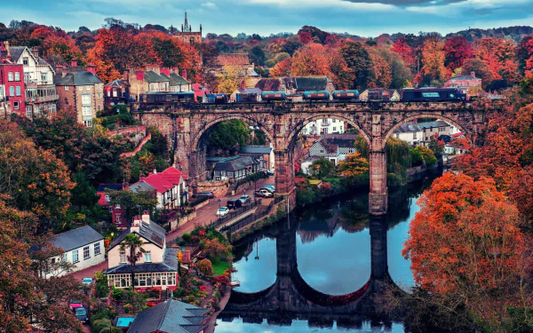 A vibrant fall scene in a Yorkshire town, featuring a train crossing a historic stone bridge over a river, surrounded by colorful autumn trees and charming houses in England.