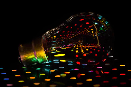 A man-made light bulb lies on a surface with colorful, vivid dots reflected inside and around it, captured in 4K Ultra HD for a striking desktop wallpaper.