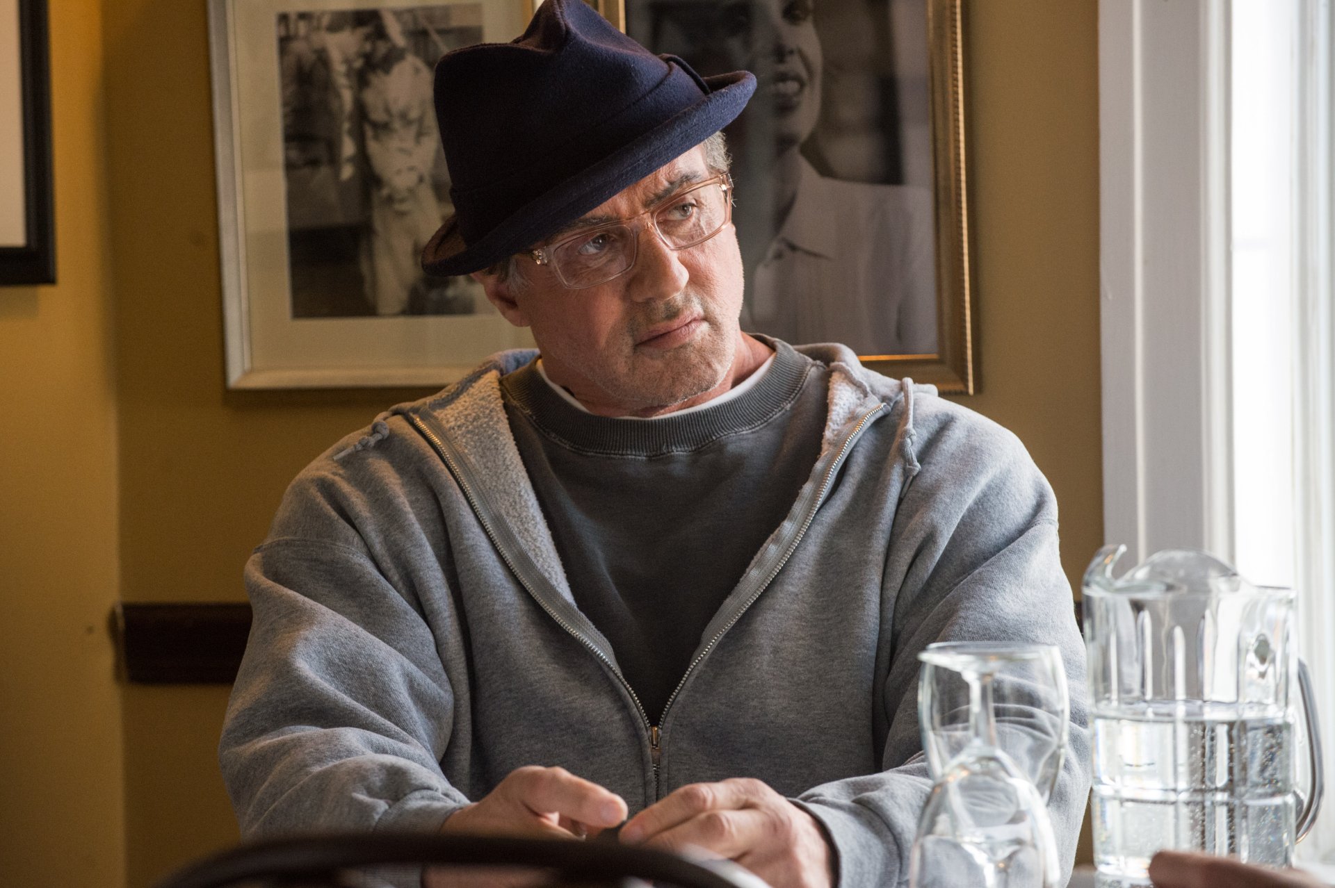 4K Ultra HD desktop wallpaper: older male film character in a hat and hoodie seated at a table, cinematic still from the Creed/Rocky boxing film franchise.