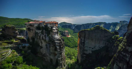 A breathtaking 4K Ultra HD view of a monastery perched atop a steep cliff in Meteora, Greece, surrounded by lush mountains under a clear blue sky.