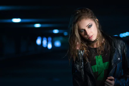 A brunette woman with blue eyes poses in a dark, urban setting. She wears a leather jacket and a green graphic t-shirt, showcasing her model presence in a striking HD wallpaper.
