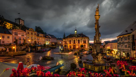 5K Ultra HD PC desktop wallpaper: Night view of an Austrian town square with illuminated historic buildings and a tall ornate man-made fountain beneath stormy clouds.