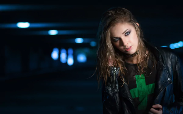 A brunette woman with blue eyes poses in a dark, urban setting. She wears a leather jacket and a green graphic t-shirt, showcasing her model presence in a striking HD wallpaper.