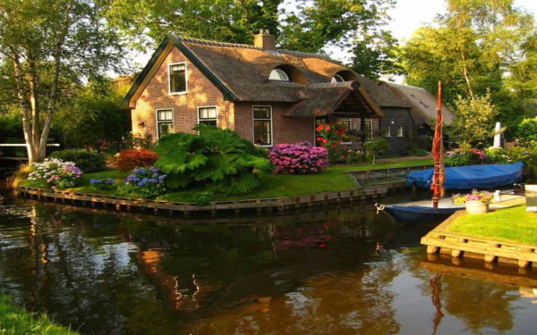 A serene spring scene featuring a thatched-roof house surrounded by blooming flowers and bushes beside a calm canal with a docked boat.