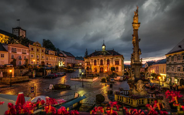 5K Ultra HD PC desktop wallpaper: Night view of an Austrian town square with illuminated historic buildings and a tall ornate man-made fountain beneath stormy clouds.