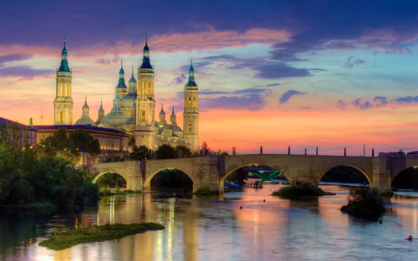 Sunset view of the Cathedral-Basilica of Our Lady of the Pillar and a bridge over the river in a Spanish city, with colorful skies reflecting on the water.