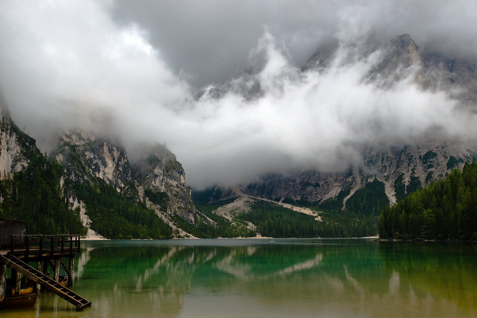 HD PC desktop wallpaper showing a serene mountain landscape with low-hanging clouds over a calm green lake surrounded by dense forest.