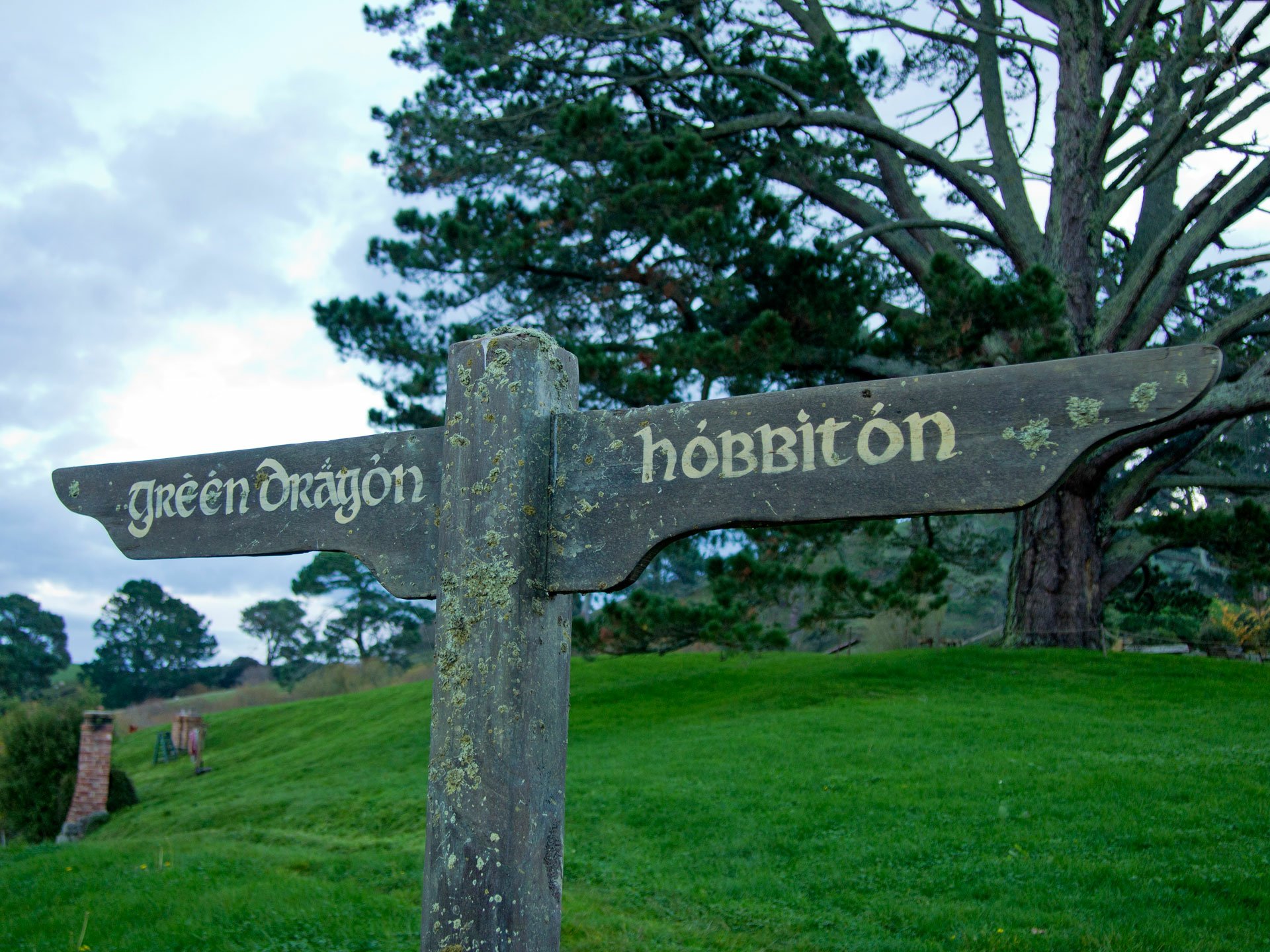 Man-made Hobbiton signpost reading Green Dragon and Hobbiton against lush rolling hills and trees — HD PC desktop wallpaper background.