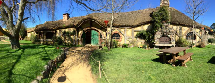 5K Ultra HD PC desktop wallpaper: man-made Hobbiton set — sunlit thatched cottage with a round green door, winding path and grassy lawn beneath a bright blue sky.