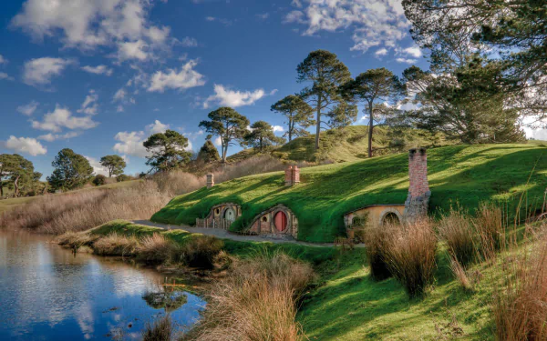 HD desktop wallpaper of Hobbiton’s man-made hobbit holes nestled in lush green hills beside a reflective pond under a partly cloudy blue sky.