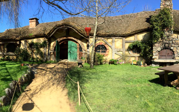 5K Ultra HD PC desktop wallpaper: man-made Hobbiton set — sunlit thatched cottage with a round green door, winding path and grassy lawn beneath a bright blue sky.