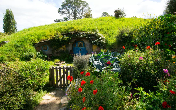 4K Ultra HD wallpaper of a man-made Hobbiton scene featuring a charming green hill hobbit hole surrounded by vibrant flowers and lush greenery under a bright sky.