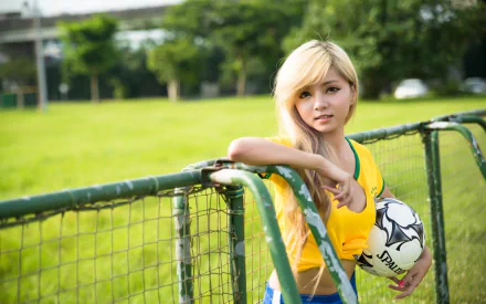 An Asian woman in a yellow sports jersey holds a soccer ball, leaning against a fence on a green field. The image is a high-definition desktop wallpaper and background.