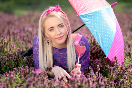 A blonde woman with long hair and bright lipstick lies in a purple flower field, holding a pink and blue polka-dot umbrella and a heart-shaped lollipop in this 4K Ultra HD wallpaper.