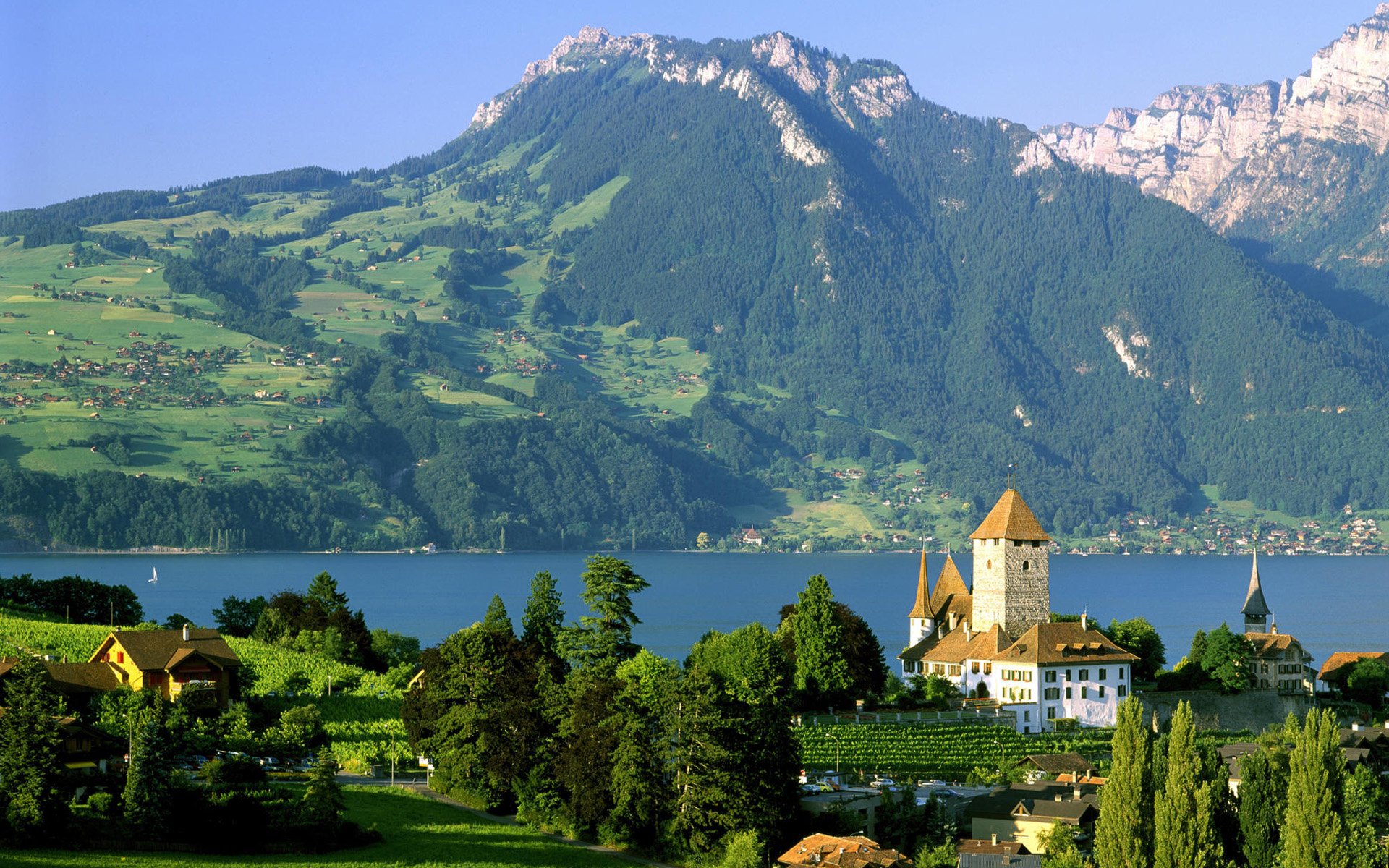 HD PC desktop wallpaper showing a vibrant landscape with a castle by a lake, lush greenery, and towering mountains under a clear blue sky.