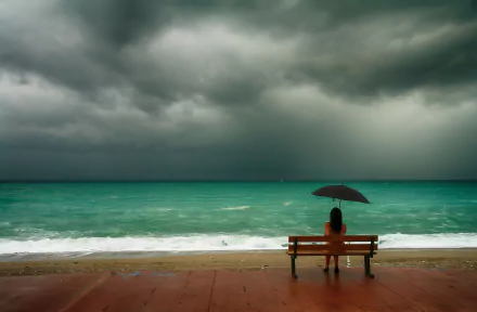 A woman sits alone on a bench by the ocean, holding an umbrella, with a cloudy horizon in the background. The mood is contemplative. This HD desktop wallpaper captures serenity and solitude.