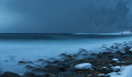 HD desktop wallpaper showing a foggy coastline with smooth stones along the shore, ocean waves, and a distant horizon blending into misty blue mountains.