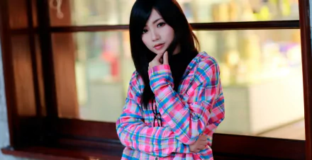 HD wallpaper featuring an Asian brunette model with brown eyes, wearing a colorful plaid shirt, posing thoughtfully in front of a blurred indoor background.